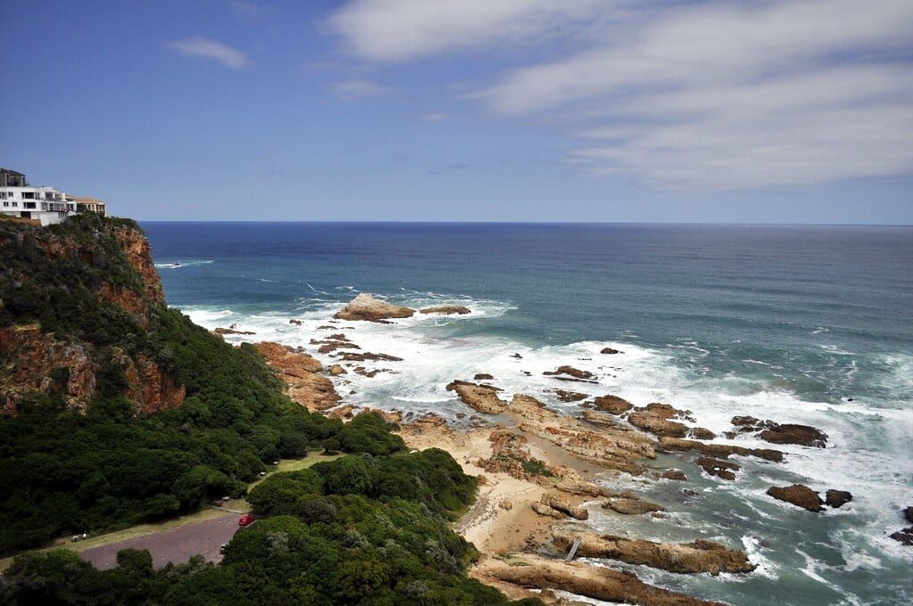 Rugged green cliffs and coastal homes overlooking waves breaking on the rocky shores of the Garden Route near Knysna.
