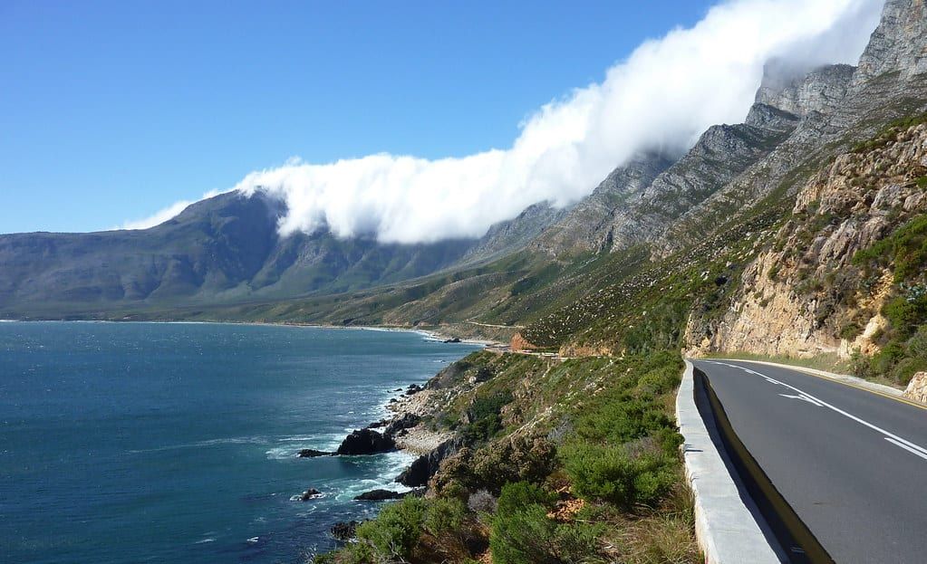 A coastal highway curves along rocky cliffs beside the ocean toward cloud-covered mountains on the Garden Route in South Africa.