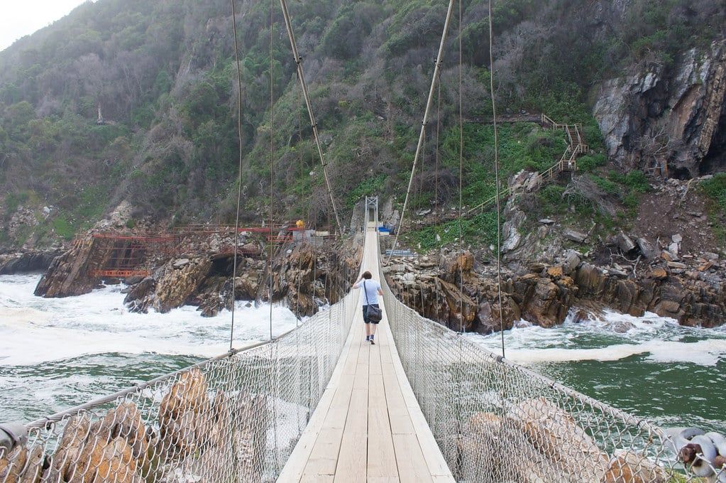 Ein Besucher geht über die Holz-Hängebrücke über die tosenden Wasser der Storms River Mouth entlang der Garden Route.