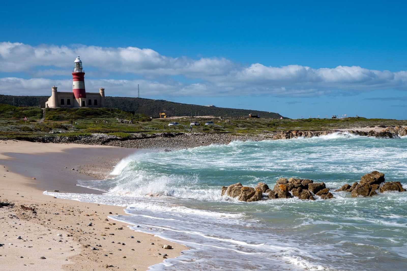 The red-and-white striped Cape Agulhas lighthouse stands on a coastal hill behind a sandy beach and crashing waves.