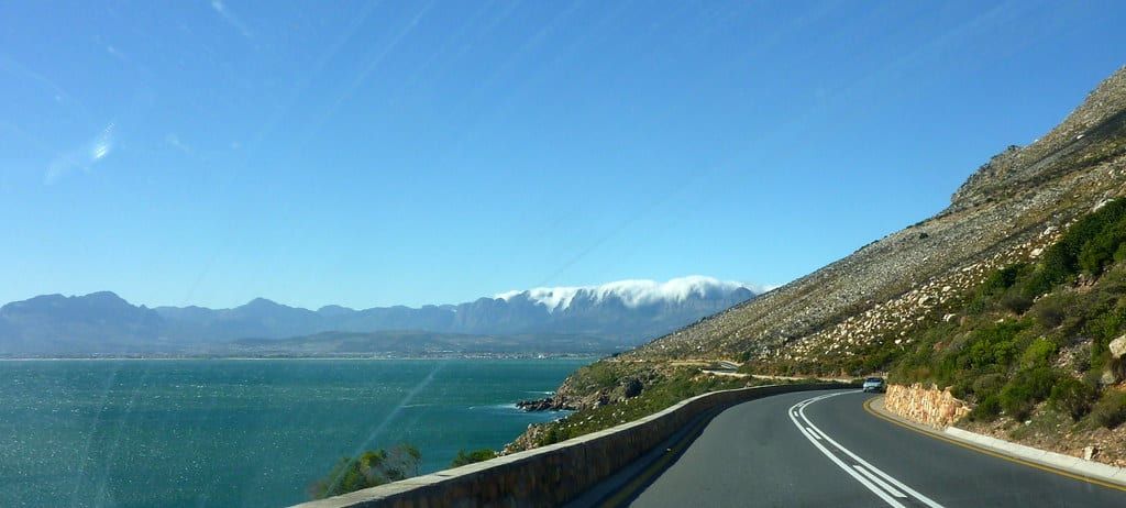 Eine asphaltierte Küstenstraße schlängelt sich entlang der Garden Route zwischen schroffen Bergen und dem türkisfarbenen Ozean unter strahlend blauem Himmel.